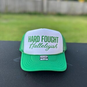 May include: Green and white trucker hat with the text "HARD FOUGHT Hallelujah" in green lettering. The hat has a green mesh back and a black label that says "OTTO TRUCKER".