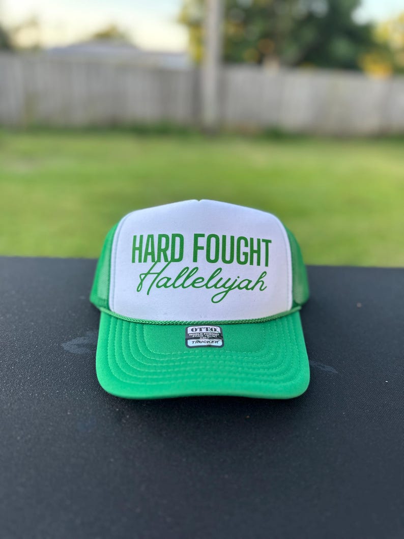 May include: Green and white trucker hat with the text "HARD FOUGHT Hallelujah" in green lettering. The hat has a green mesh back and a black label that says "OTTO TRUCKER".