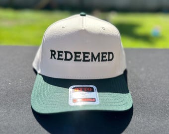 May include: A beige baseball cap with a dark green brim and the word REDEEMED embroidered in black capital letters on the front panel. The cap features a small green button on top and a rectangular sticker on the brim with the brand name OTTO.