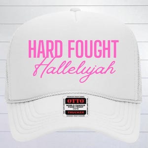 May include: White trucker hat with a pink embroidered design that says "Hard Fought Hallelujah".