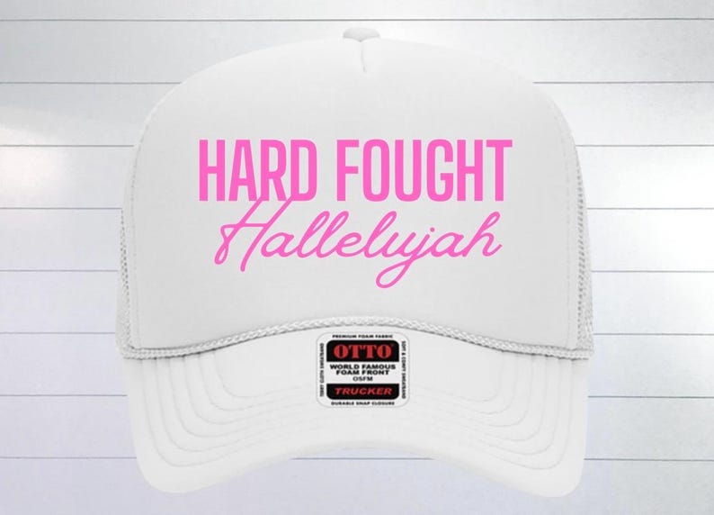 May include: White trucker hat with a pink embroidered design that says "Hard Fought Hallelujah".