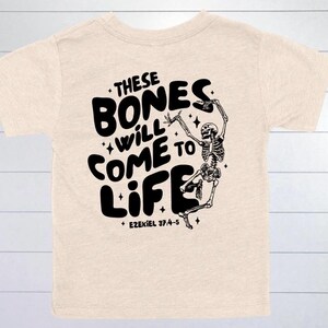 May include: Beige t-shirt with a black graphic of a dancing skeleton and the words "THESE BONES WILL COME TO LIFE" in a bold, playful font. The text "Ezekiel 37:4-5" is printed below the phrase.