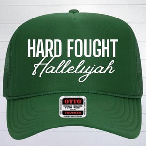 May include: Green trucker hat with a white embroidered design that says "Hard Fought Hallelujah". The hat has a black mesh back and a black label that says "Otto World Famous Foam Front Caps Trucker".