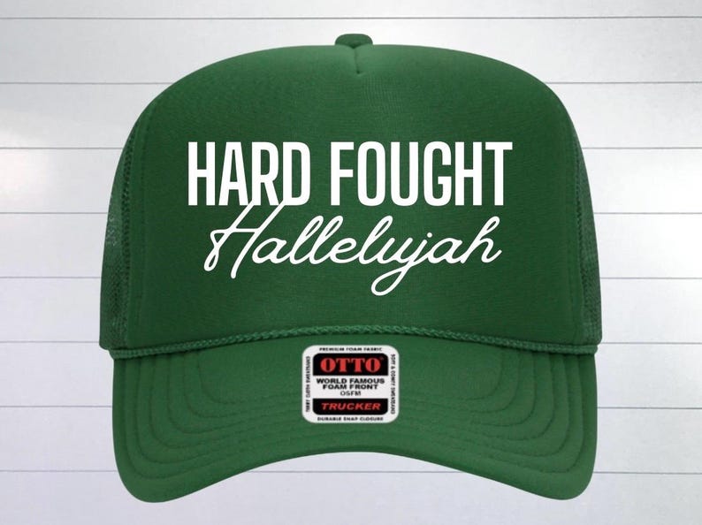 May include: Green trucker hat with a white embroidered design that says "Hard Fought Hallelujah". The hat has a black mesh back and a black label that says "Otto World Famous Foam Front Caps Trucker".