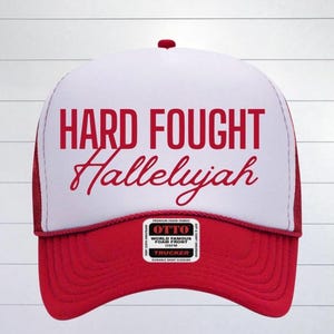 May include: A red and white trucker hat with the text "HARD FOUGHT Hallelujah" in red lettering. The hat has a red mesh back and a red bill.