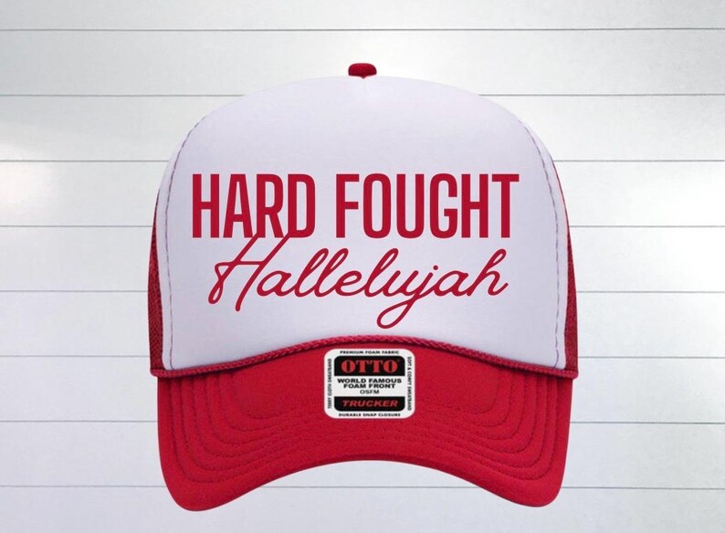 May include: A red and white trucker hat with the text "HARD FOUGHT Hallelujah" in red lettering. The hat has a red mesh back and a red bill.