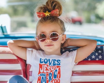 Puede incluir: Una persona joven con gafas de sol redondas y una camiseta blanca con el texto "Loves Jesus and America Too". Tiene un lazo rojo con una estrella dorada en el pelo. El fondo incluye el interior de un coche con un diseño de bandera americana.