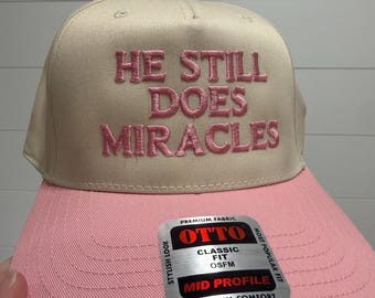 May include: A beige baseball cap with a pink brim and pink embroidered text on the front that reads HE STILL DOES MIRACLES. A silver and black sticker on the brim displays the text OTTO PREMIUM FABRIC CLASSIC FIT OSFM MID PROFILE.
