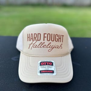 May include: A beige trucker hat with a brown embroidered design that says "HARD FOUGHT Hallelujah". The hat has a black mesh back and a brown adjustable strap.