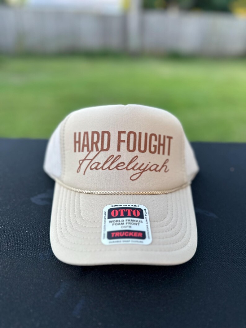 May include: A beige trucker hat with a brown embroidered design that says "HARD FOUGHT Hallelujah". The hat has a black mesh back and a brown adjustable strap.