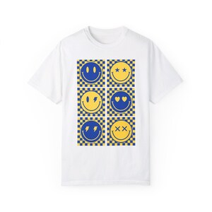 Peut inclure: T-shirt blanc avec une grille de six visages souriants. Chaque visage souriant a une expression différente et est placé sur un fond à carreaux jaune et bleu. Les visages souriants sont dans des tons de jaune et de bleu.