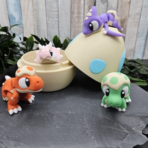 Könnte beinhalten: Ein Set mit vier kleinen Dinosaurier-Figuren, darunter ein lilafarbener Dinosaurier auf einem beigefarbenen Ei, ein rosafarbener Dinosaurier in einem beigefarbenen Ei, ein orangefarbener Dinosaurier und ein grüner Dinosaurier mit grünen Flecken.