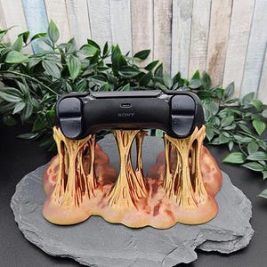Könnte beinhalten: Ein schwarzer Sony PlayStation-Controller liegt auf einem einzigartigen Ständer. Der Ständer hat ein strukturiertes, organisches Design in Gold- und Rotbrauntönen, das von säulenartigen Strukturen getragen wird. Der Controller und der Ständer sind auf einem grauen, unregelmäßig geformten Stein platziert.
