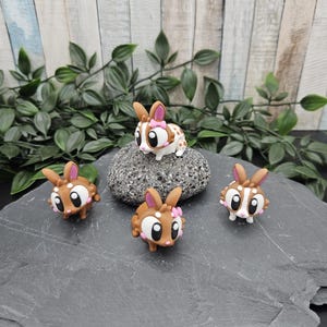 Könnte beinhalten: Eine Sammlung kleiner, dekorativer Hasenfiguren. Die Häschen sind braun und weiß mit rosa Akzenten, einschließlich Schleifen. Sie sind auf einer grauen Steinoberfläche mit Grünzeug im Hintergrund angeordnet. Die Hasen haben große, ausdrucksstarke Augen.