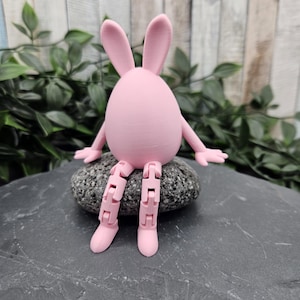 Könnte beinhalten: Eine rosa, eiförmige Hasenfigur mit beweglichen Beinen und Armen, die auf einem grauen Stein sitzt. Der Hase hat lange Ohren und ist mit ausgestreckten Armen posiert. Der Hintergrund zeigt Grün und eine Holzoberfläche.