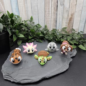 Könnte beinhalten: Fünf kleine, bunte, cartoonartige Tierfiguren. Die Figuren sind ein braunes und schwarzes Gecko, ein rosa und gelbes Axolotl, ein grauer Seehund, eine grüne Schildkröte und ein brauner und weißer Hund mit einem rosa Halsband.