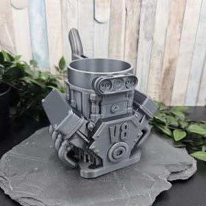 Könnte beinhalten: Ein silberfarbener Becher in Form eines V8-Motorblocks. Der Becher zeigt detaillierte mechanische Komponenten, darunter Zylinderköpfe, Abgaskrümmer und ein markantes V8-Emblem auf der Vorderseite. Er hat eine zylindrische Öffnung an der Oberseite.