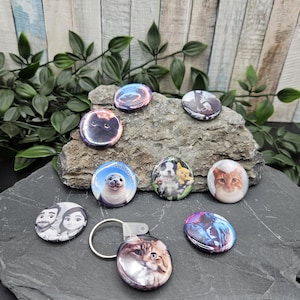 Könnte beinhalten: Eine Sammlung runder Ansteckbuttons und ein Schlüsselanhänger mit verschiedenen Tier- und Porträtmotiven. Die Buttons zeigen Bilder einer Katze, einer Robbe, eines Hundes und eines Paares. Der Schlüsselanhänger hat ein Katzenbild. Die Artikel werden auf einer dunkelgrauen Oberfläche präsentiert.