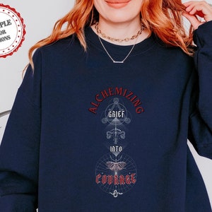 May include: Navy blue sweatshirt with a graphic design featuring a geometric pattern and the text "Alchemizing Grief into Courage".