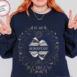 Puede incluir: Sudadera azul marino con un gráfico dorado de un libro con el texto "In My Romantasy Era" rodeado de estrellas, lunas y otros símbolos celestiales.
