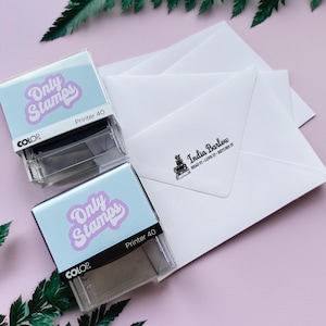 May include: Two clear plastic self-inking stamps with a light blue background and the text "Only Stamps" in a purple retro font. The text "Printer 40" is printed in black below the logo. The stamps are sitting on a pink surface.