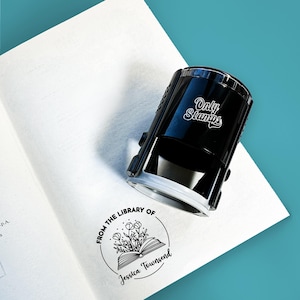 Op de afbeelding: Een zwarte zelf-inktende stempel met de tekst "Only Stamps" aan de zijkant. De stempel wordt gebruikt om een ronde, zwarte inkt stempel te maken met de tekst "From the Library of Jessica Townsend" en een tekening van een open boek met bloemen.