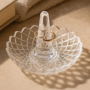 Könnte beinhalten: Ein klarer Glasringhalter mit einem filigranen, geometrischen Design. Der Halter verfügt über einen erhöhten Mittelpfosten zum Präsentieren eines Rings.