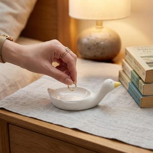 May include: A white ceramic duck-shaped ring holder with a yellow beak. The ring holder is on a light grey linen surface, next to a stack of books and a lamp. A gold ring is being placed in the holder.
