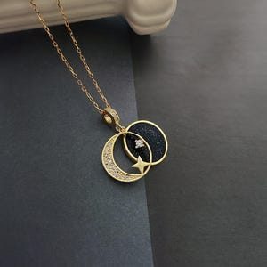 Può includere: Una collana con catena dorata con un ciondolo a forma di luna crescente e stella. La luna è nera con una texture scintillante e un piccolo ciottolo trasparente al centro. La stella è dorata con un ciottolo trasparente al centro.