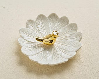 May include: White ceramic ring dish with a gold bird design. The dish is shaped like a fan of feathers.