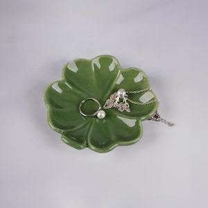 Puede incluir: Un plato de cerámica verde en forma de trébol con un anillo y un collar de plata en la parte superior.