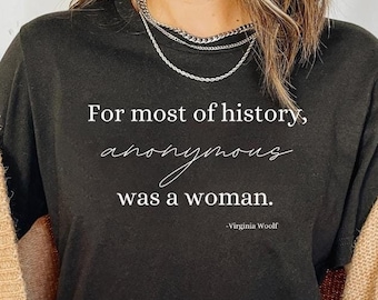 Puede incluir: Camiseta negra con la cita "For most of history, anonymous was a woman." en texto blanco, atribuida a Virginia Woolf. La camiseta se combina con una rebeca beige y vaqueros azules.