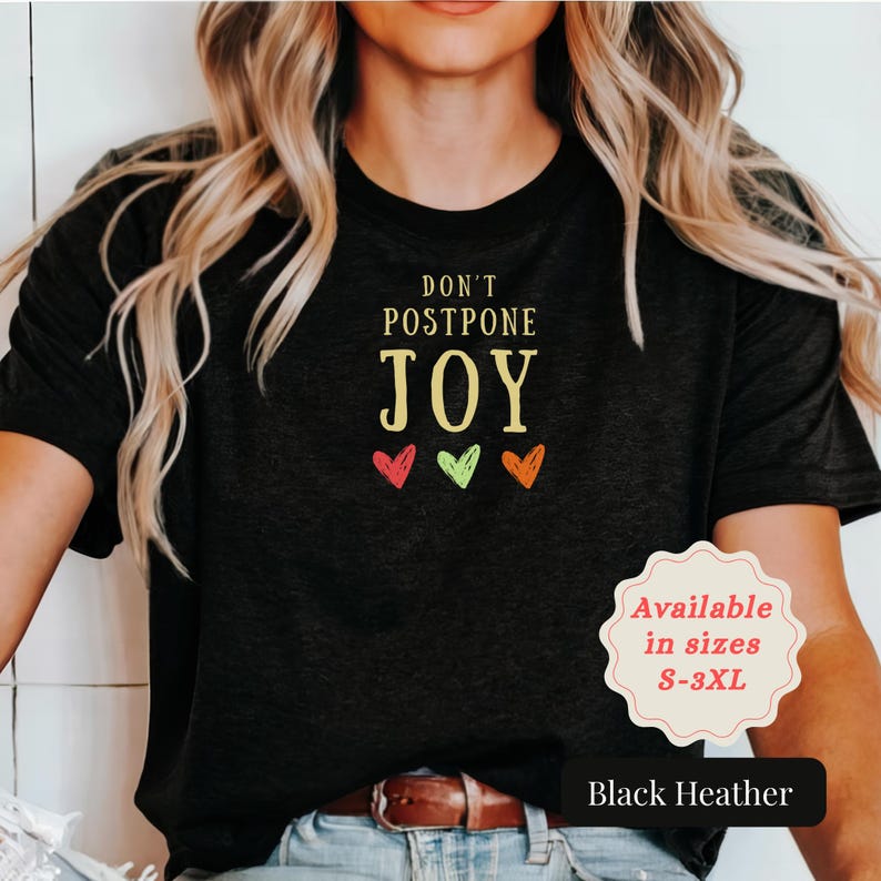 May include: A black heather t-shirt featuring the text DON'T POSTPONE JOY in yellow lettering, accompanied by three small hand-drawn style hearts in red, light green, and orange colors centered below the text on the chest area.