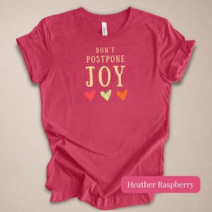 May include: A heather raspberry short-sleeve t-shirt featuring the text DON'T POSTPONE JOY in a light yellow font, centered above three small hand-drawn style hearts in red, light green, and orange colors, with a crew neckline and a standard fit.