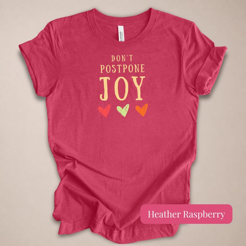 May include: A heather raspberry short-sleeve t-shirt featuring the text DON'T POSTPONE JOY in a light yellow font, centered above three small hand-drawn style hearts in red, light green, and orange colors, with a crew neckline and a standard fit.