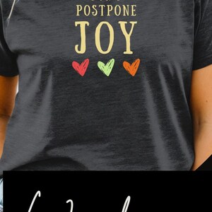 May include: A collection of three short-sleeve t-shirts. The first is blue with the text "doing my best-ish". The second is black with the text "DON'T POSTPONE JOY" and three small hearts. The third is cream with the text "be the good."