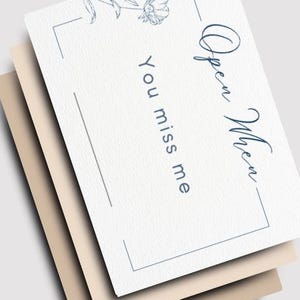 May include: Three cards stacked on top of each other. The top card is white with the words "Open When" and "You miss me" in a handwritten font. A floral illustration and a thin blue border decorate the card. The other two cards are beige and tan.