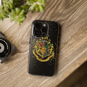 May include: Black smartphone case featuring the Hogwarts crest in gold, red, green, and blue. The case has a glossy finish and a speckled design. The phone case is on a wooden surface.