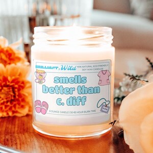 May include: A scented candle in a glass jar with the text "Smells better than C. diff" on a label. A pair of black wick trimmers and orange flowers are in the foreground. A rose and a coffee cup are in the background.