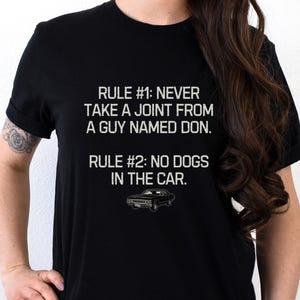 Pode incluir: Uma t-shirt preta de manga curta com texto branco que diz: RULE #1: NEVER TAKE A JOINT FROM A GUY NAMED DON. RULE #2: NO DOGS IN THE CAR. Abaixo do texto, há uma pequena e simples ilustração gráfica de um carro preto.