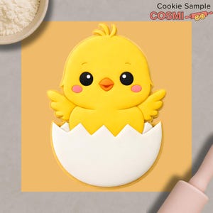 Op de afbeelding: Een gele kuikenkoekjessnijder die uit een witte eierschaal komt. Het kuiken heeft zwarte ogen, een oranje snavel en roze wangen. De afbeelding bevat de tekst "Cookie Sample" en "COSMI" met een icoon van een deegroller.