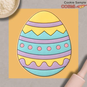 Puede incluir: Una colorida galleta con forma de huevo de Pascua con secciones amarillas, celeste y moradas. El huevo está decorado con patrones en zigzag y ondulados, y puntos rosas. La galleta está sobre un fondo amarillo con el texto "Cookie Sample" y la marca "COSMI".