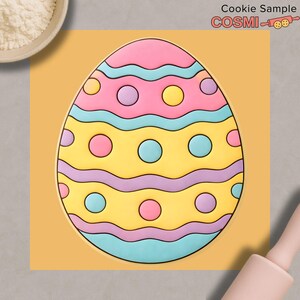 Puede incluir: Una galleta decorada con forma de huevo de Pascua con glaseado rosa, amarillo, azul y morado. La galleta en forma de huevo tiene un patrón ondulado y detalles circulares. La imagen también incluye un cuenco de harina, un rodillo y el texto "Cookie Sample COSMIC".