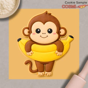 May include: A brown monkey cookie with a yellow banana, smiling with rosy cheeks. The cookie is on a yellow background. The text "Cookie Sample" and "COSMI" are in the upper right corner.