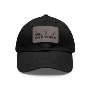 May include: A black baseball cap featuring a rectangular grey patch on the front. The patch displays black icons of a lawnmower, two spatulas, and a fishing rod, with the text DAD VIBES printed in bold black capital letters below the icons.