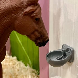 May include: A brown horse figurine positioned next to a small, grey, wall-mounted water bowl. The bowl is semi-circular with a central lever mechanism, designed to be attached to a vertical surface, and features a textured, matte finish.