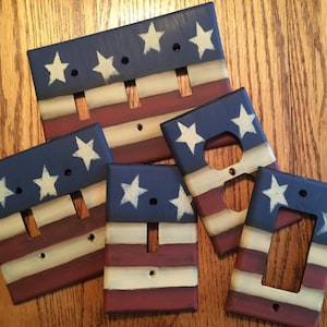 May include: Five wooden light switch and outlet covers painted with an American flag design. The covers are painted in shades of blue, red, and white with white stars on a blue background.
