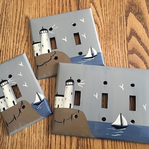 May include: Three light switch covers painted with a nautical theme. Each cover features a lighthouse, a sailboat, and seagulls against a blue and gray background.