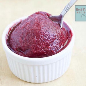 May include: A white bowl filled with a vibrant red, smooth, and creamy fruit puree. The puree is thick and appears to be made from berries. A spoon is partially submerged in the bowl, suggesting the puree is ready to be enjoyed.