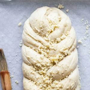 Può includere: Un filone di pane intrecciato ricoperto di briciole bianche su una superficie bianca. Il pane ha la forma di una treccia ed è di colore marrone chiaro. Le briciole sono sparse intorno al pane e sulla superficie. Il testo "Real Food Recipes" è visibile nell'angolo in alto a destra dell'immagine.